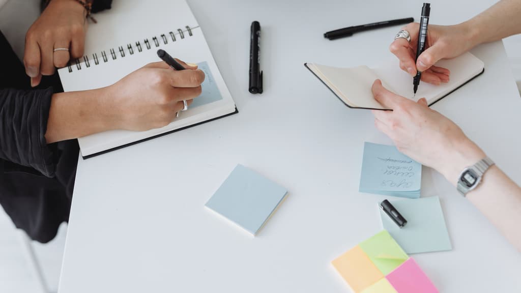 Hands writing in notebooks among sticky notes on a white table